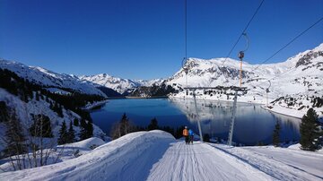 A snow-covered landscape with a clear lake and mountains in the background. Two people are standing on the slope and enjoying the view.