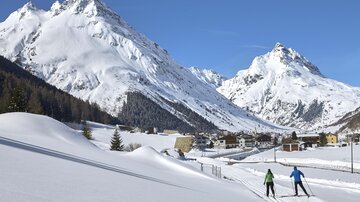 Two skiers glide through a snowy landscape. In the background, an impressive mountain scenery rises under a clear blue sky.