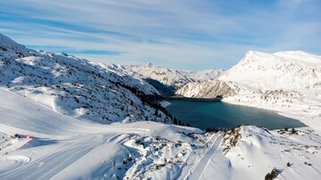 A wintry landscape with snow-covered mountains and a clear, blue lake. The sky is bright and cloudless.