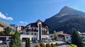 A picturesque alpine landscape with a traditional hotel building and majestic mountains in the background. The sky is clear, and the surroundings are green and inviting.