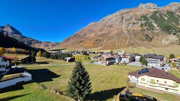 A picturesque mountain landscape with a clear blue sky and autumn-colored meadows. In the foreground, cozy houses and a single tree can be seen.
