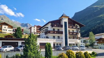 A beautiful hotel in the Alps with a modern façade and surrounded by mountains. The surroundings are green and inviting.