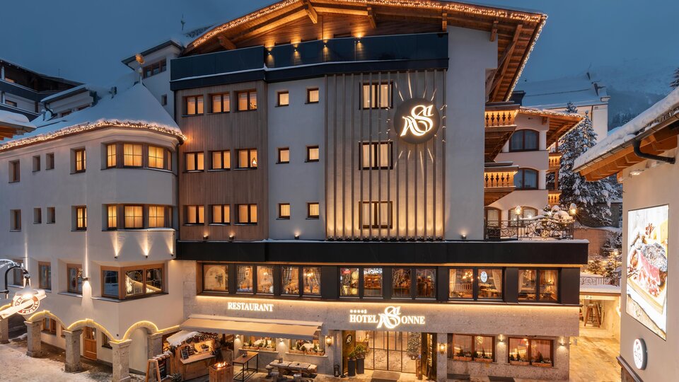 A beautiful hotel building in alpine style, festively illuminated at dusk. Below it is a restaurant with an inviting outdoor area.