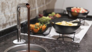 A stainless steel chocolate fountain, from which liquid chocolate flows, stands on a table. In the background, there are fruit bowls with various fruits like apples and grapes.