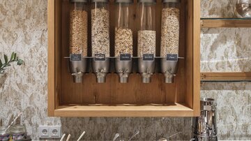 A breakfast bar with many different mueslis in dispenser systems. Additionally, fresh fruits and snacks are arranged on a table.