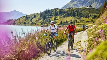 Two cyclists ride along a beautiful forest path by a lake. The surroundings are green and hilly, with colorful flowers by the roadside.