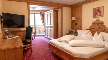 A cozy hotel room with wooden furniture and a large bed. There is a television, a desk, and a balcony with an outdoor view.