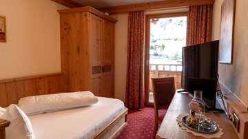 A cozy room with wooden paneling and a single bed. There is a television, a table, and a balcony with a view outside.