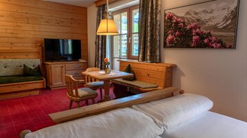 A cozy living space with wooden paneling and a small dining area. A vase of flowers sits on the window, and a picture of a mountain landscape hangs on the wall.
