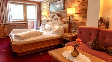 A cozy hotel room with a large double bed and a sofa. The room is decorated with wooden paneling and a carpet and offers a view of the mountains.