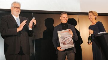 An award ceremony with three people on stage. The award winner holds a trophy and smiles, while the others applaud.